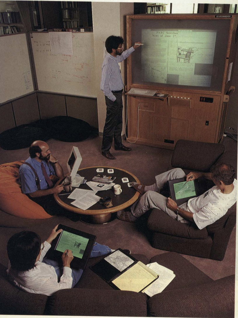 Mark Weiser at Xerox PARC pointing at a wall-sized rear-projection display showing 'PARC Scoreboard News of June 27'. Three colleagues sit on bean bags around a low table, two of them using early tablet devices that pre-date the term. The room is the PARC ubicomp lab in the early 1990s.