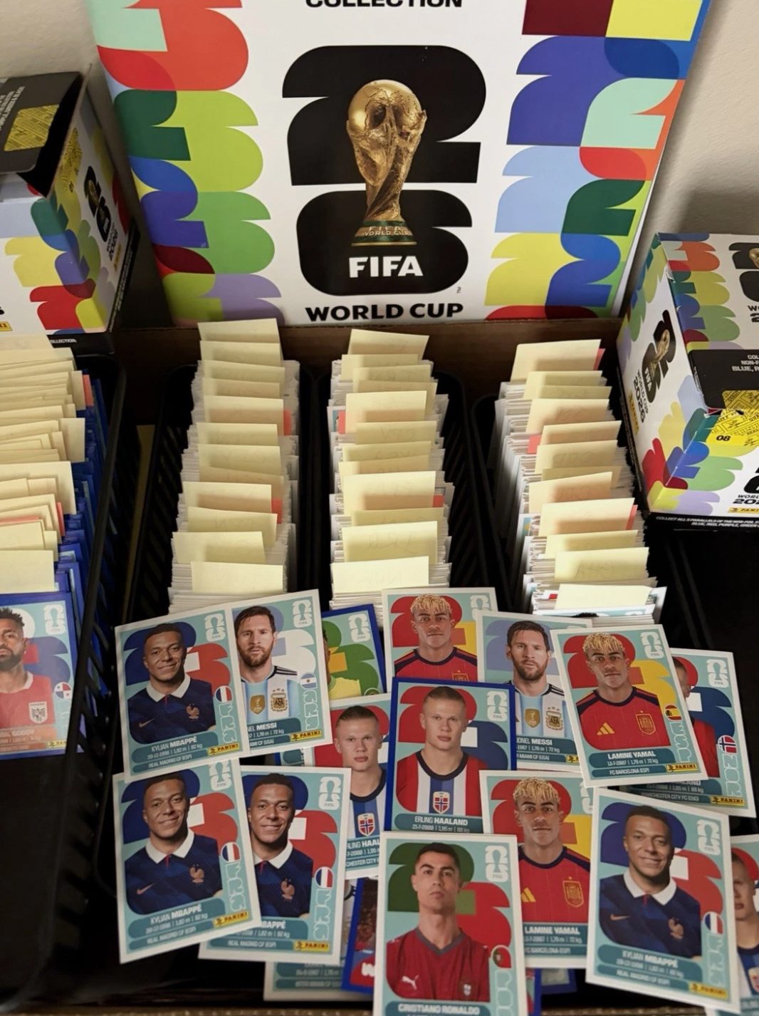 An open box of Panini 2026 World Cup stickers with rows of packets inside, and a spread of opened stickers in front showing many duplicates of Mbappé, Messi, Haaland, Yamal and Ronaldo.